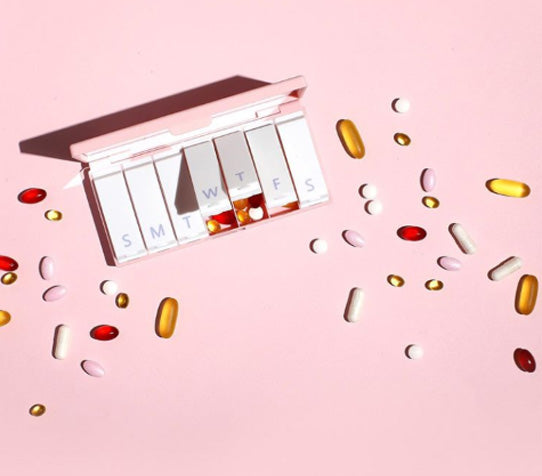 A pink Port and Polish pill box on a pink background with a variety of pills scattered throughout the photo.