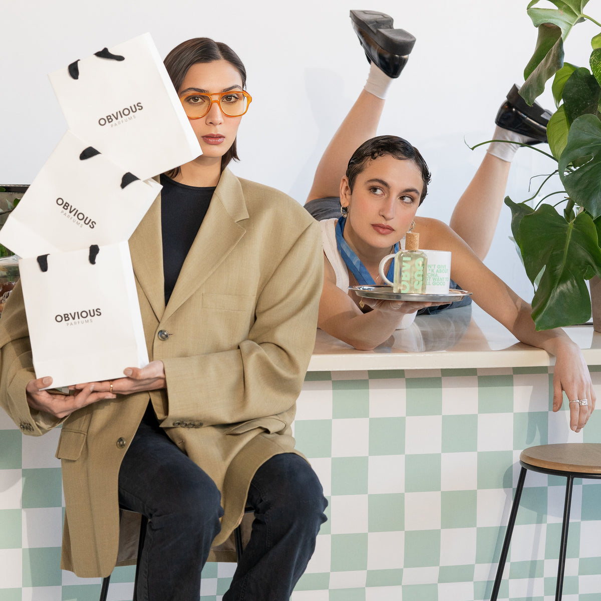 Two women in a casual setting with one holding an 'Obvious' product bag and the other leaning on a counter.