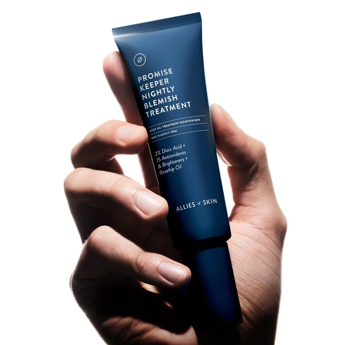 Hand holding a blue skincare tube labeled 'Promise Keeper Nightly Blemish Treatment' by Allies of Skin on a white background.
