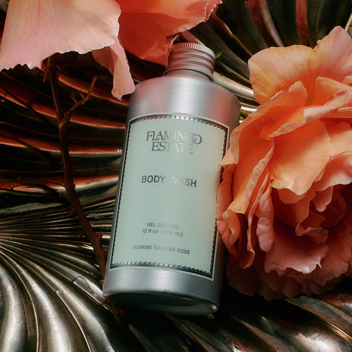 A silver bottle of Flaming Estate Body Wash, labeled Jasmine Damask Rose, styled with peach-pink flowers on a metallic leaf surface.