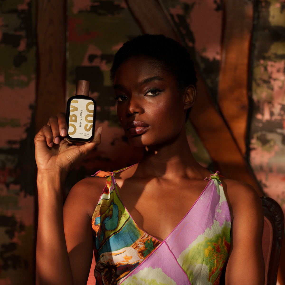 Woman holding a perfume bottle with a colorful dress sitting on a chair.