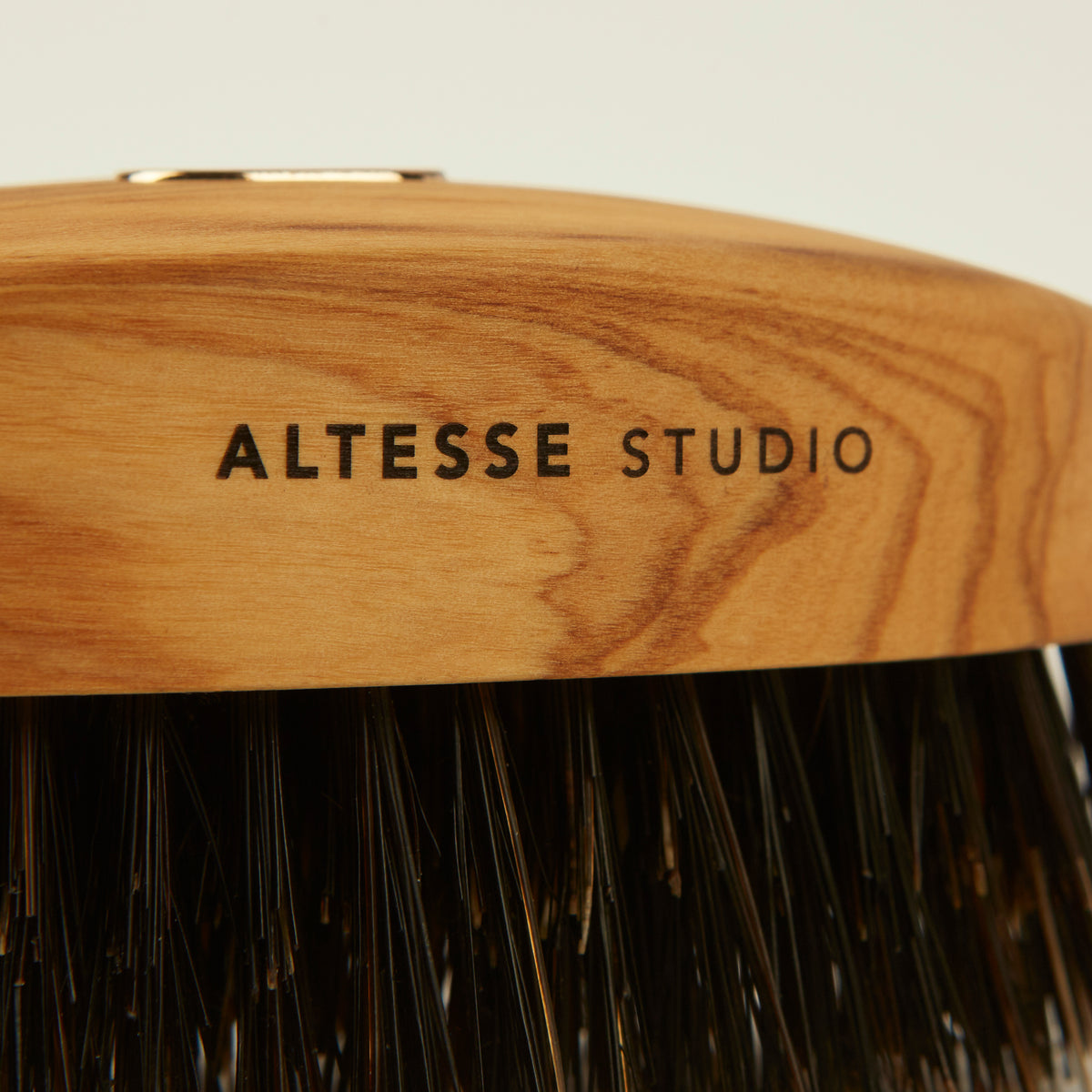 Close-up of a hairbrush with a curved natural wood handle engraved with 'ALTESSE STUDIO' and dark bristles below, on a light neutral background.