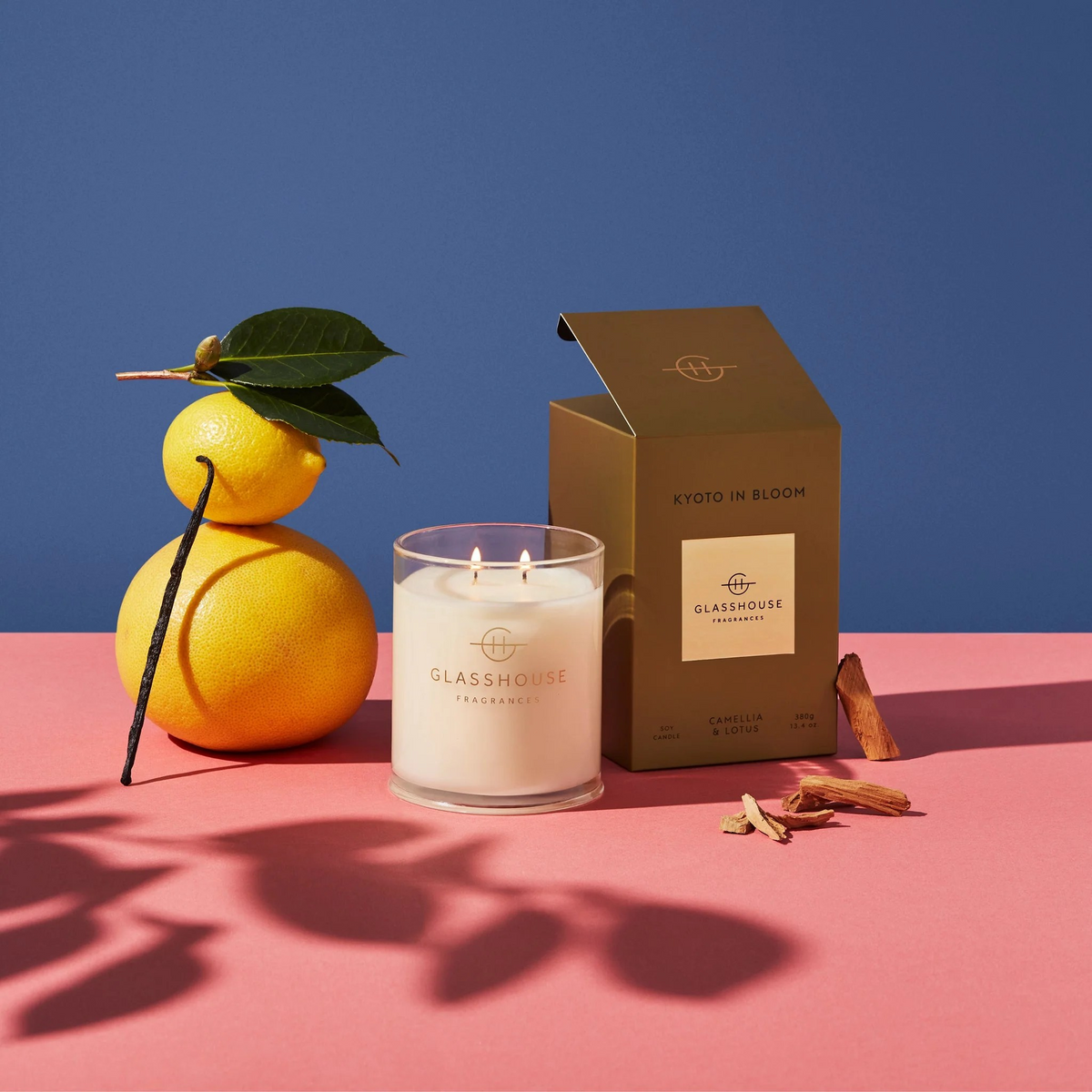 Kyoto in Bloom candle with both wicks lit with the golden brown box to the right of it and a few lemons stacked on top of each other with a a couple of spices and lemon leafs.  On a pinkish table top with a dark blue background. 