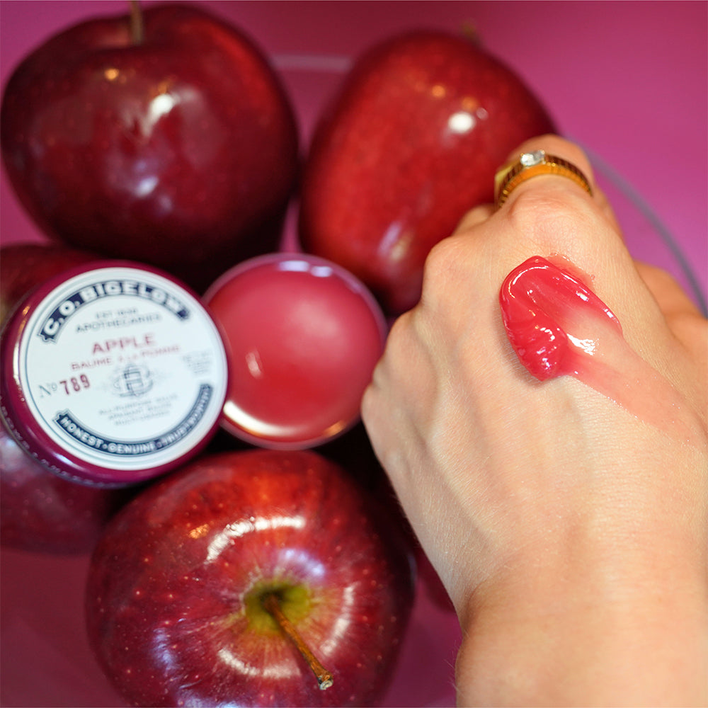 Hand swatched with C.O. Bigelow Apple All-Purpose Salve in front of an open tin of product and red apples on a burgundy background.
