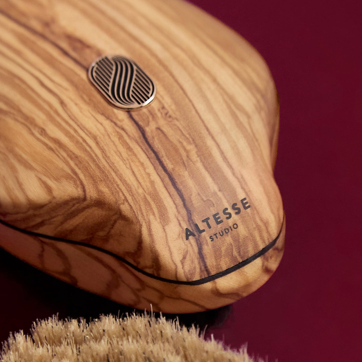 Close-up of an Altesse Studio wooden body brush with a natural olive wood grain handle, a silver metal logo emblem, and a beige bristle head, set against a dark burgundy background.