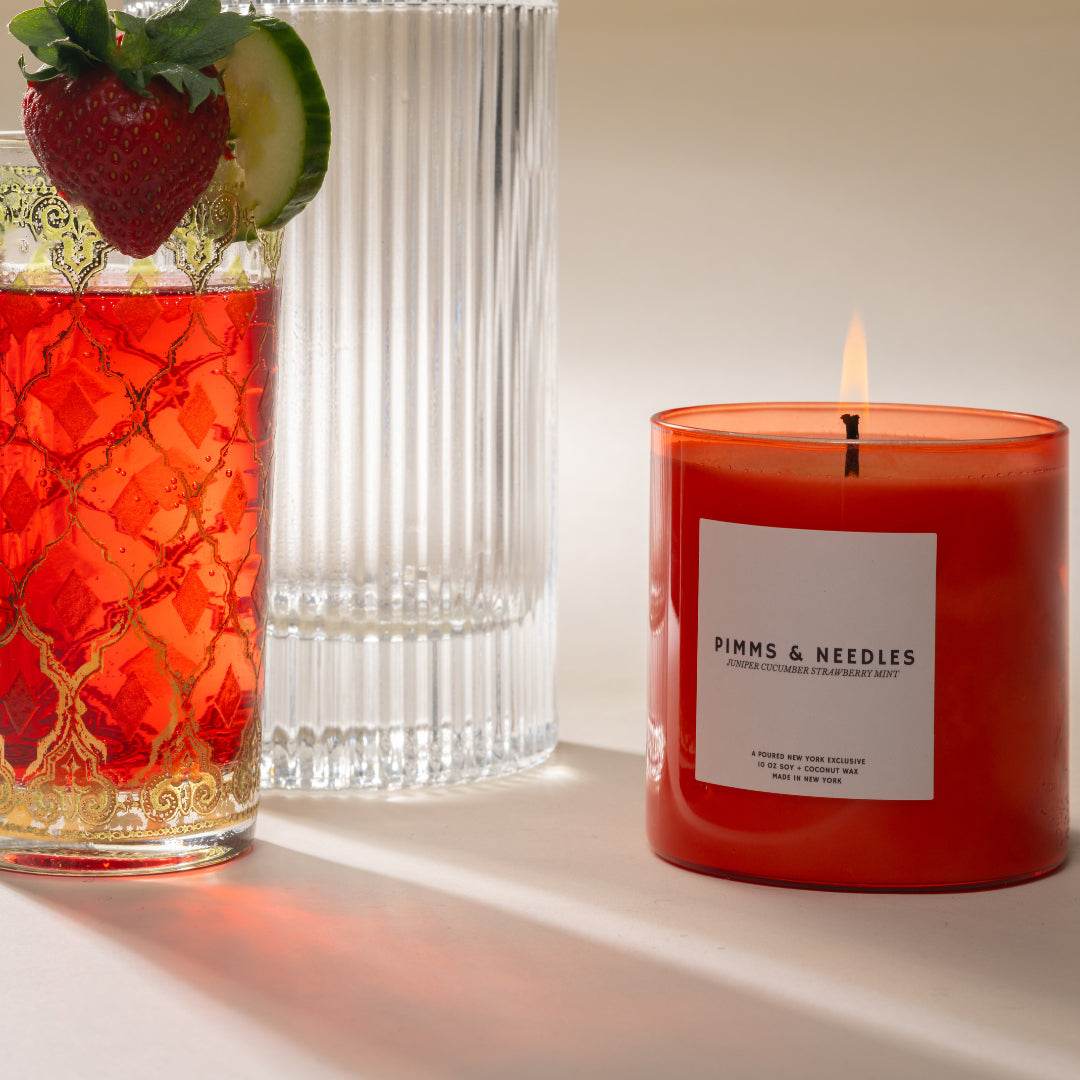 Red candle labeled 'Pimms & Needles' next to a glass of red liquid with a strawberry and lime.