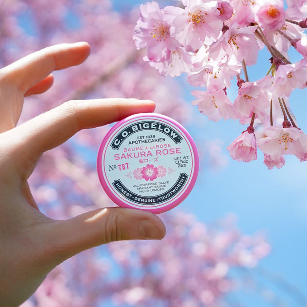 Hand holding Sakura Rose Salve Tin in front of pink flowers and a blue sky.