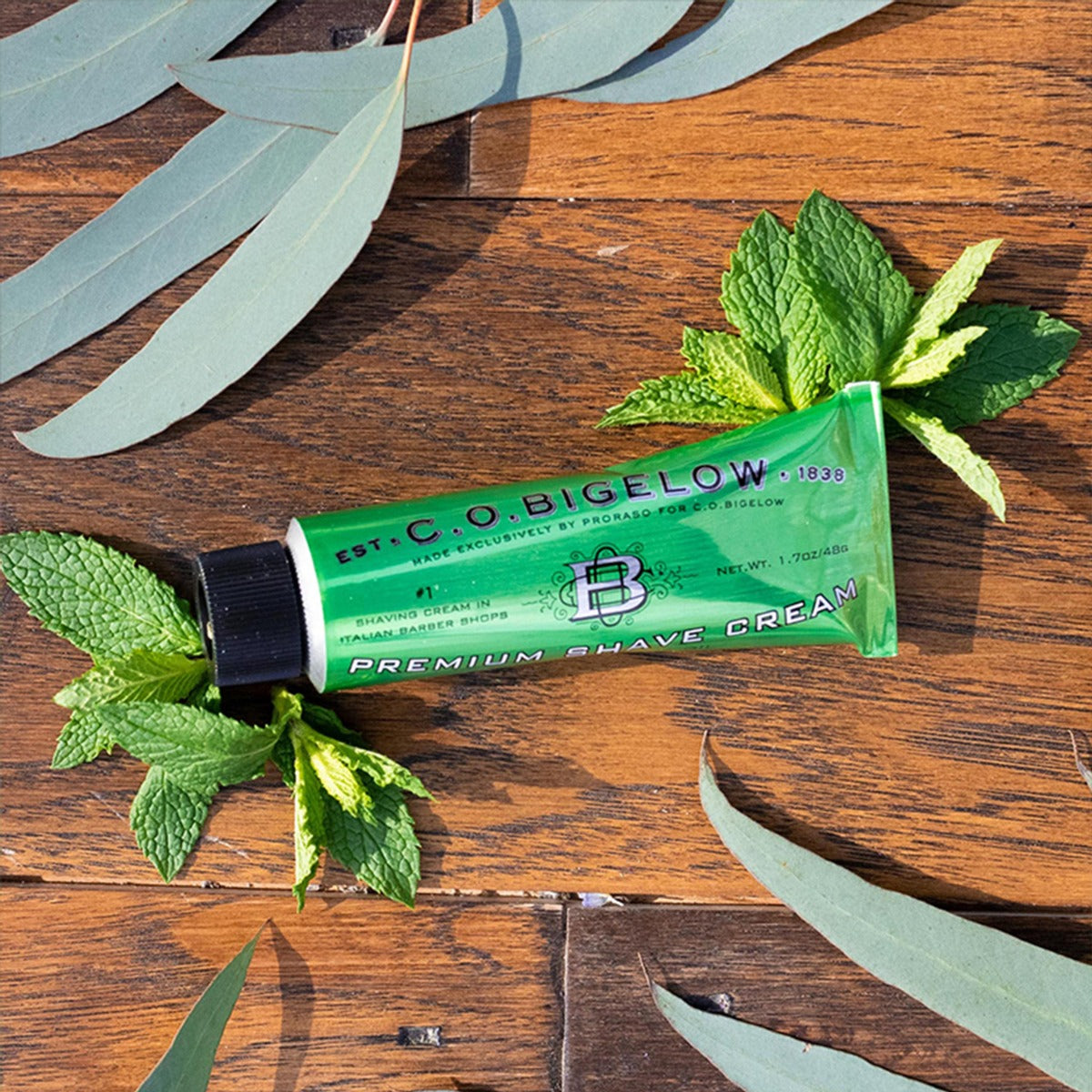 A tube of C.O. Bigelow Chemist Premium Shave Cream shown on a wooden table top with eucalyptus leaves and mint leaves around it.