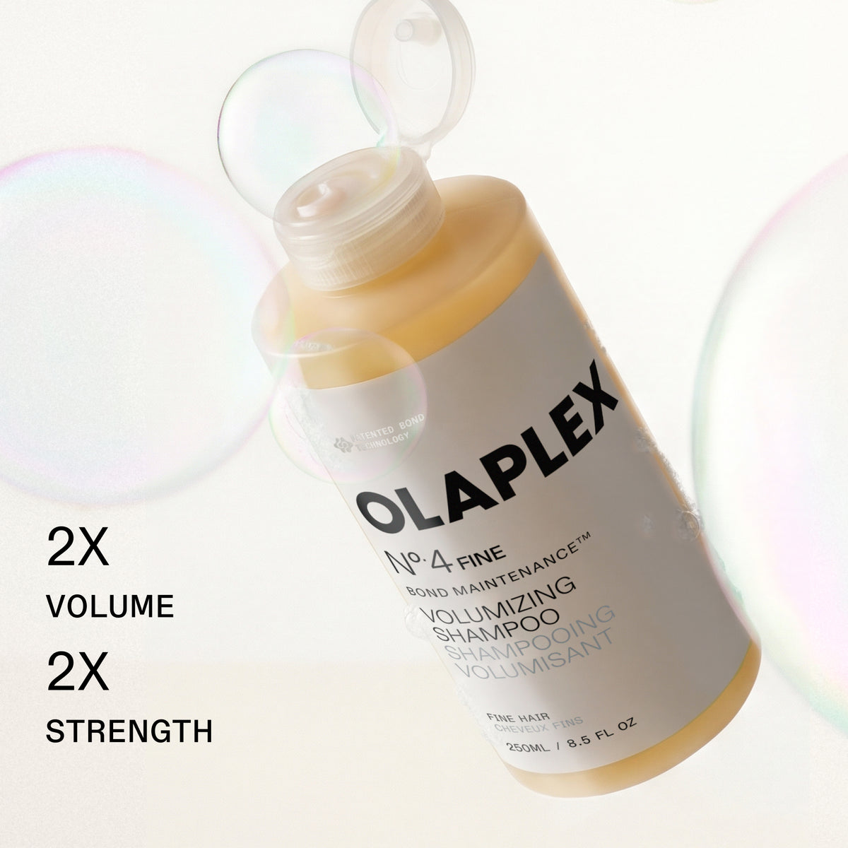 A bottle of Olaplex No. 4 Fine Volumizing Shampoo floating diagonally on a white background with iridescent soap bubbles and black text on the left reading '2X VOLUME 2X STRENGTH'.