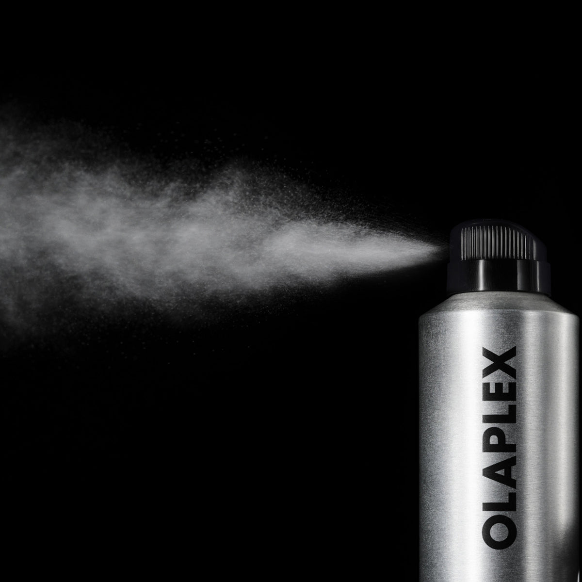 A silver aerosol spray can labeled OLAPLEX on a black background, with a fine white mist spraying from the nozzle.