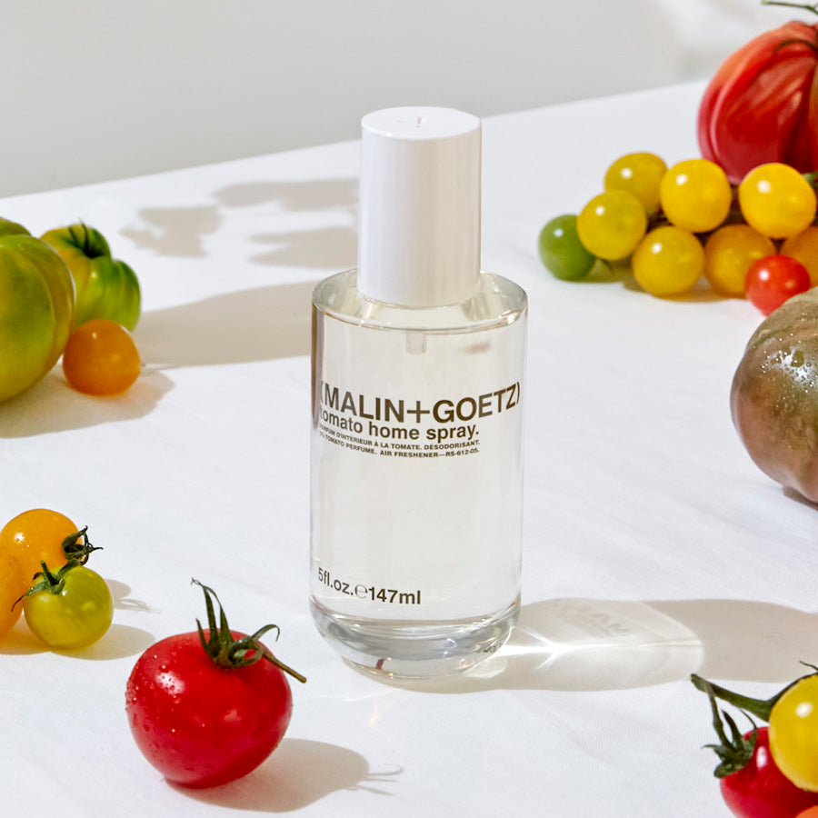 Malin+Goetz tomato home spray bottle surrounded by tomatoes and grapes on a white surface