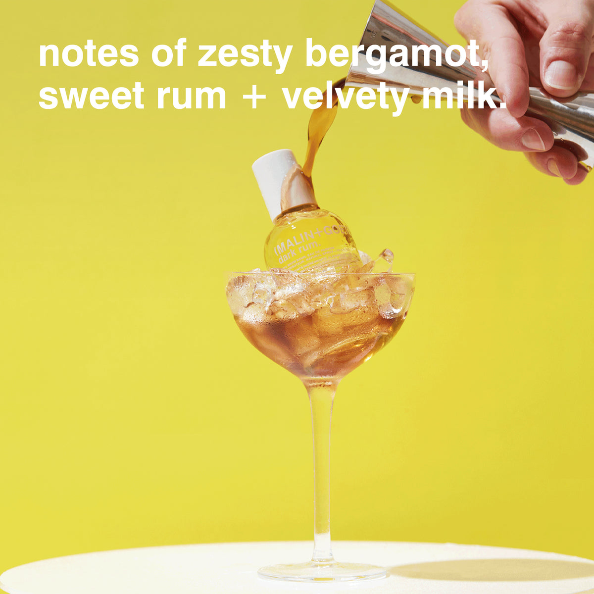 A hand pours amber liquid from a metal bottle into a stemmed glass filled with ice, set against a bright yellow background. White text at the top reads: 'notes of zesty bergamot, sweet rum + velvety milk.