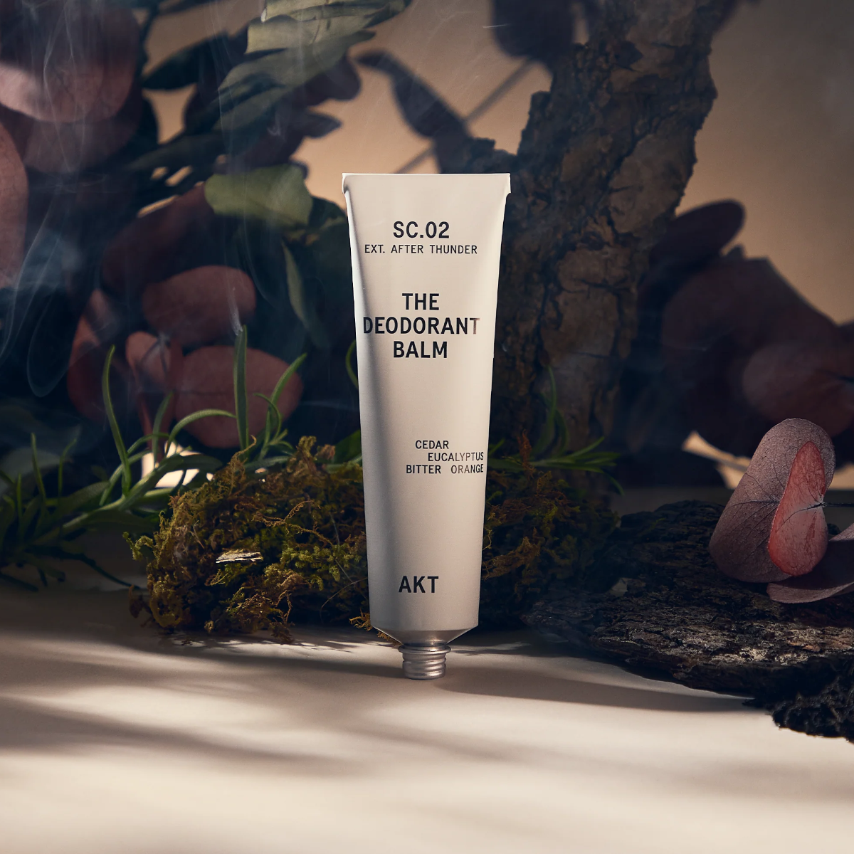 AKT After Thunder Deodorant Balm Tube standing on a gray surface in front of moss, eucalyptus leaves, and cedar wood bark. There is a smoky atmosphere around the product. 