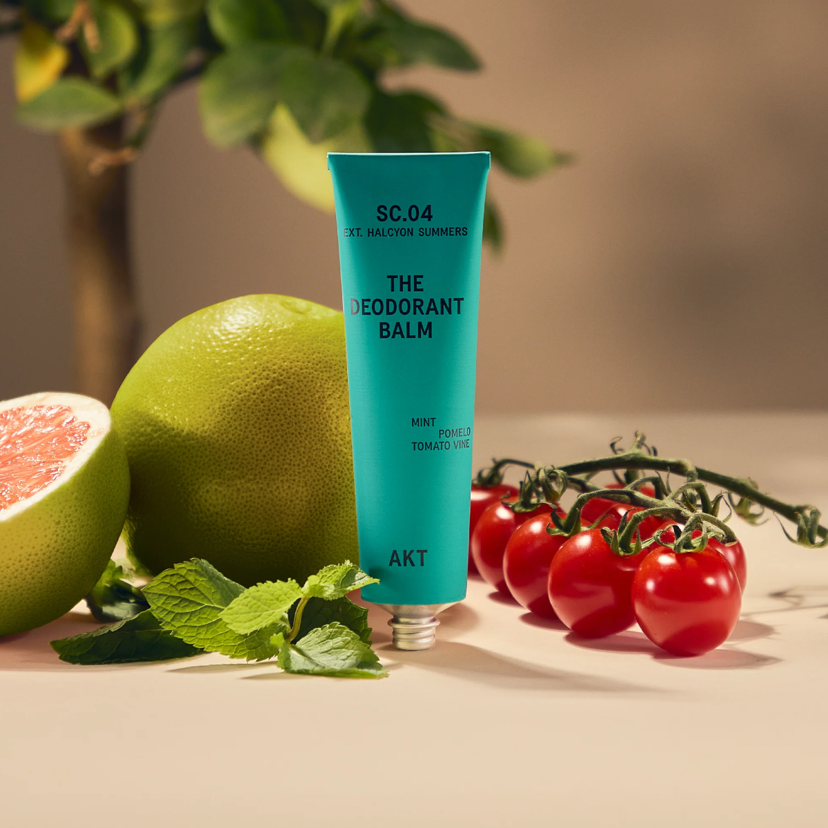 AKT Halcyon Summers Scented Deodorant Balm Tube standing on a tan surface in front of mint leaves, tomatoes, and a pomelo fruit. 