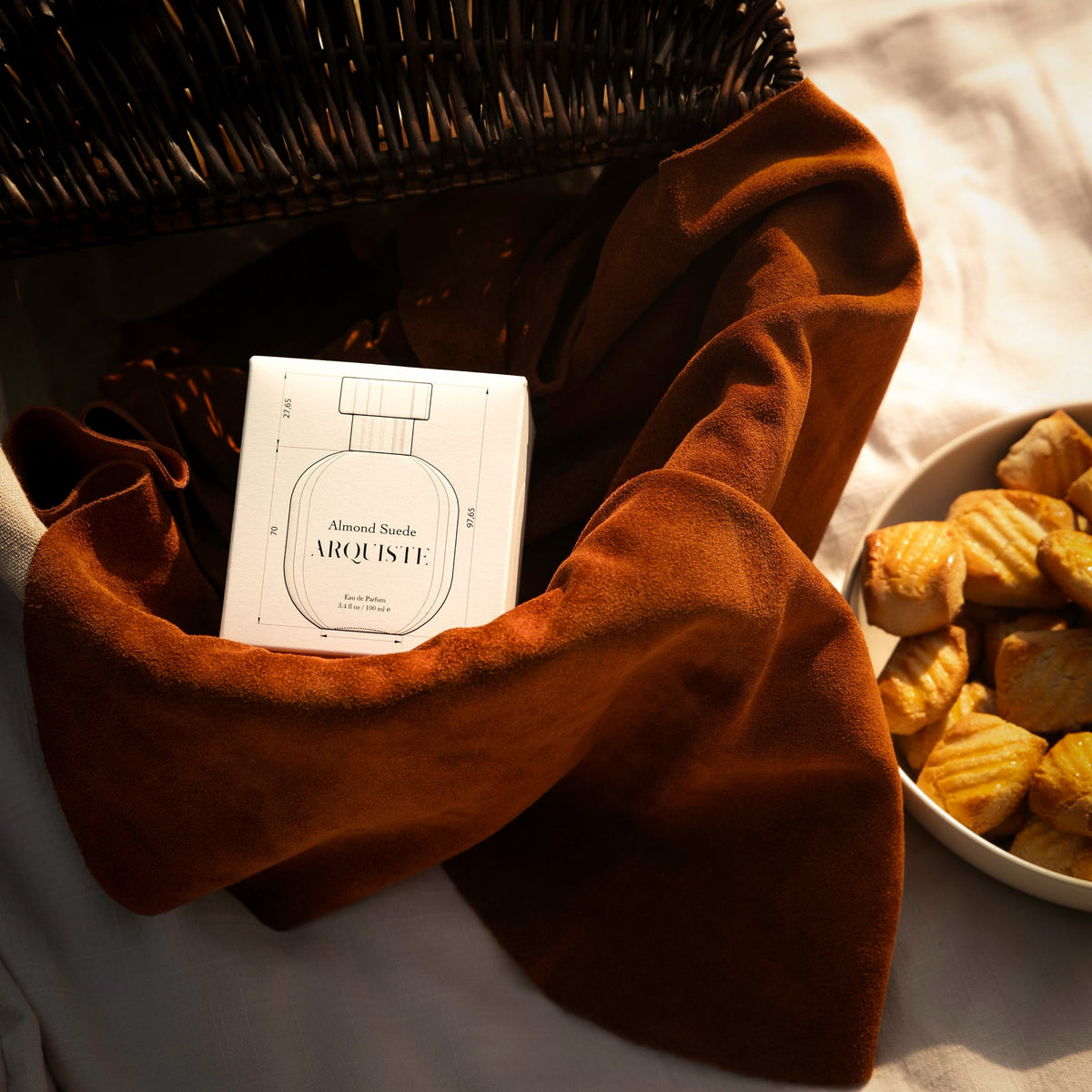 Almond Suede perfume bottle inside of a basket next to a bowl of biscuits