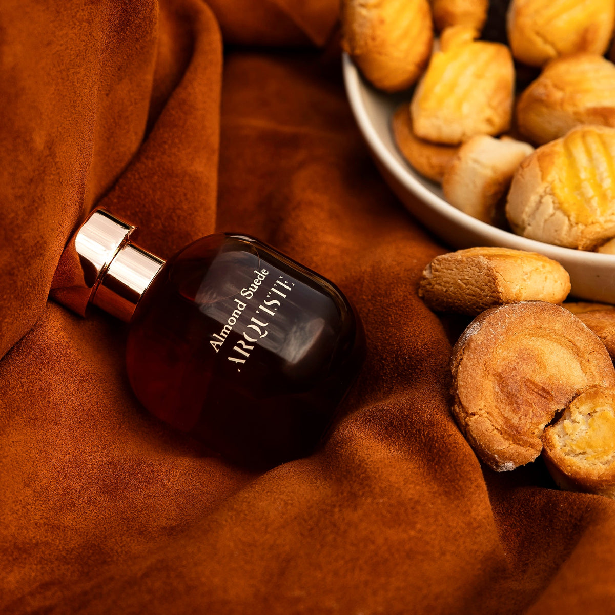 Almond Suede perfume bottle on orange suede next to a bowl of biscuits
