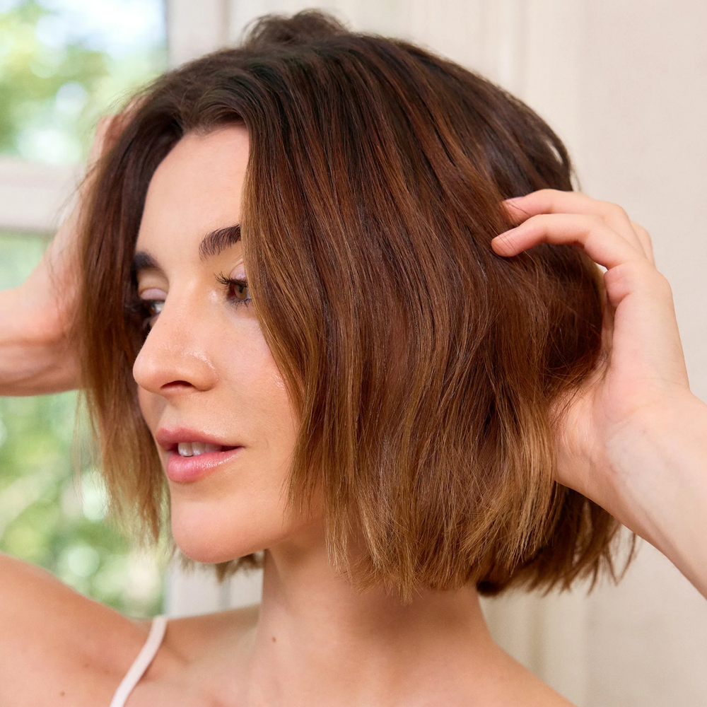 A close-up of a woman with shoulder-length brown hair, touching her hair with both hands in a bright indoor setting.