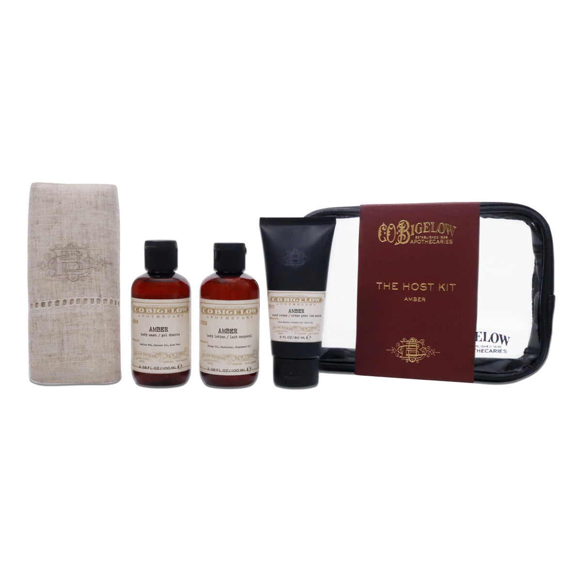 A luxury travel toiletry gift set from C.O Bigelow, featuring a beige linen towel, two amber bottles of shower and body wash, a black tube of hand cream, and a black zippered pouch with a burgundy label.
