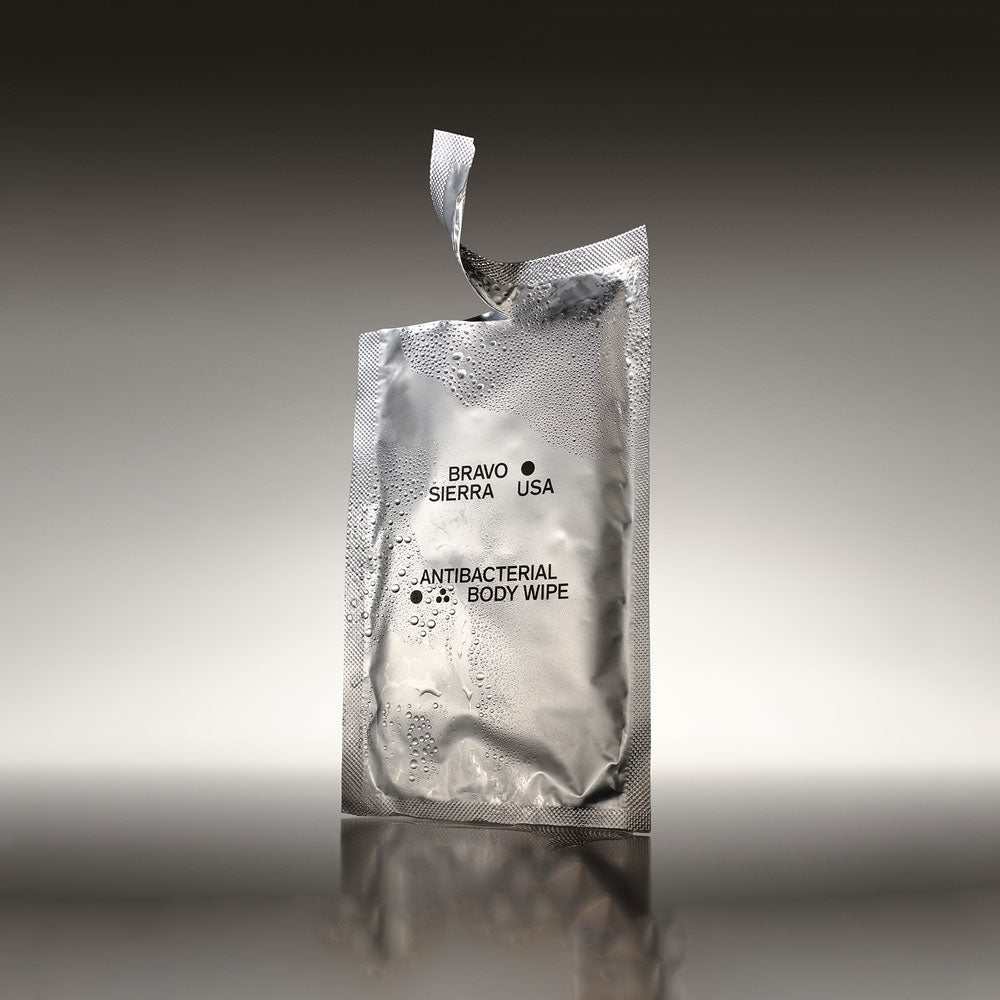 A single Bravo Sierra Antibacterial Body Wipe ripped open and covered in water droplets on top of a reflective surface in front of a white and gray background.