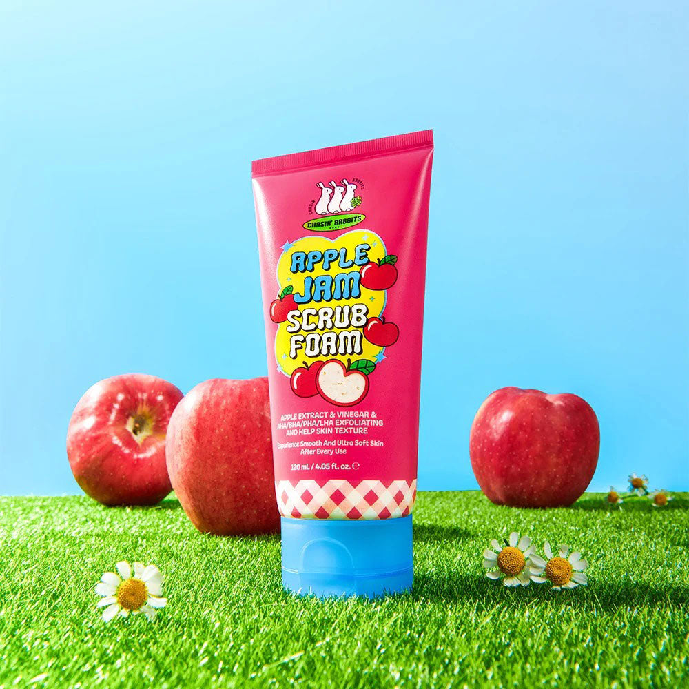 Pink tube of 'Apple Jam Scrub Foam' with apples and flowers on grass against a blue sky