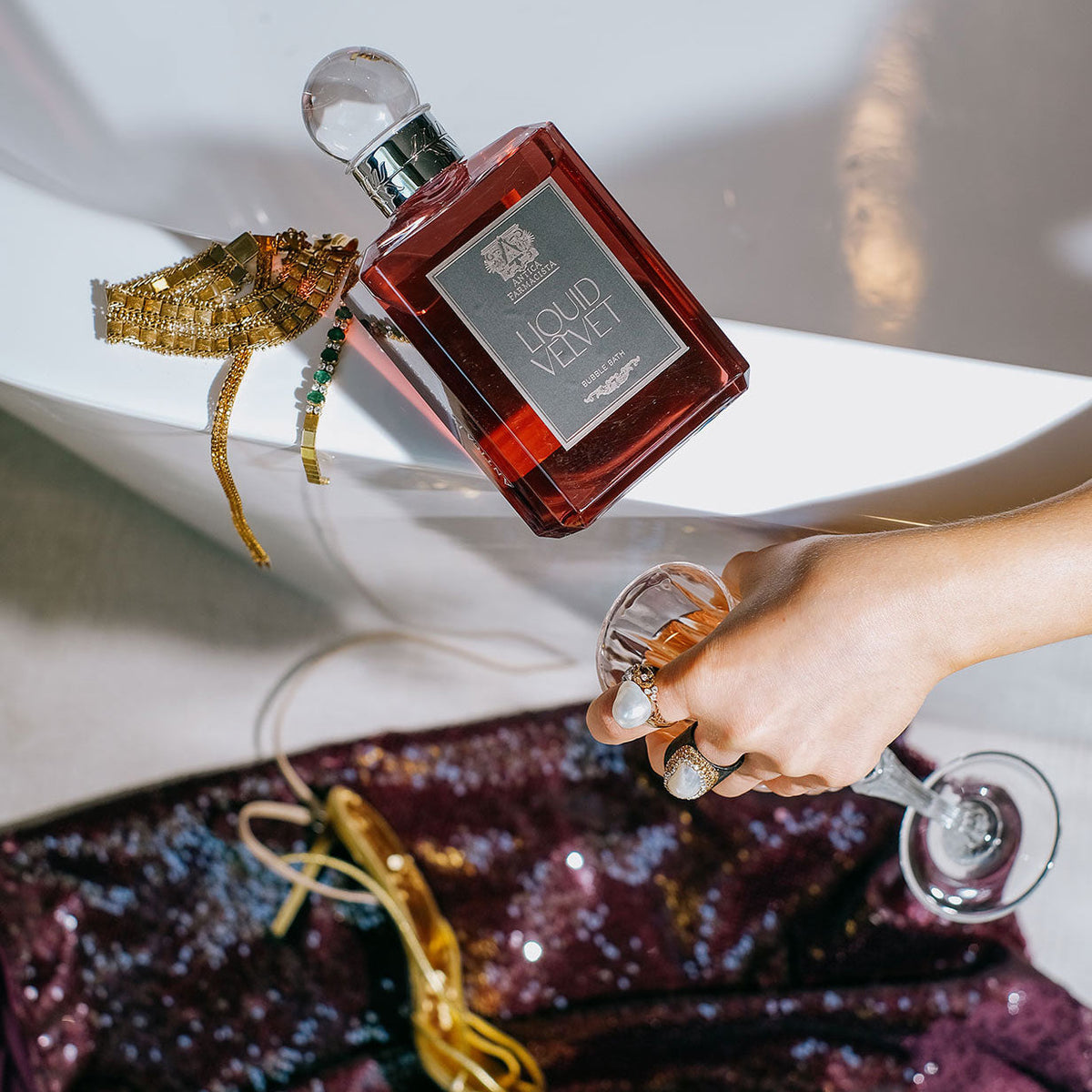 Liquid velvet bubble bath in a bottle next to gold jewelry, and a hand holding a drink, on a white and purple background