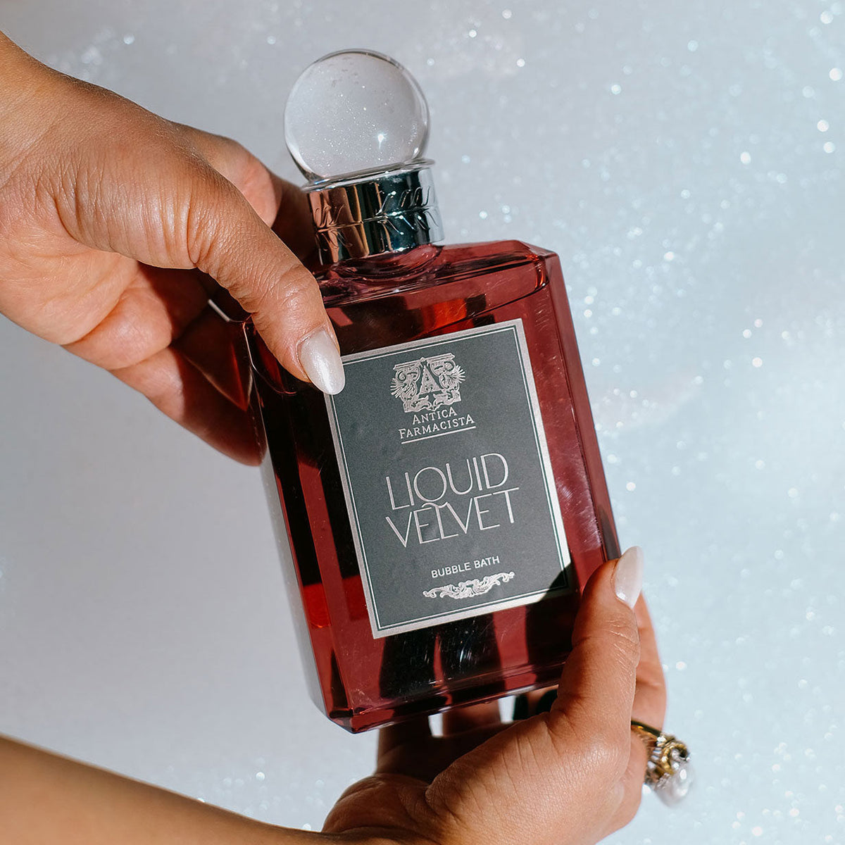 Hands Holding Liquid Velvet Bubble Bath in a bottle in front of a bubble bath