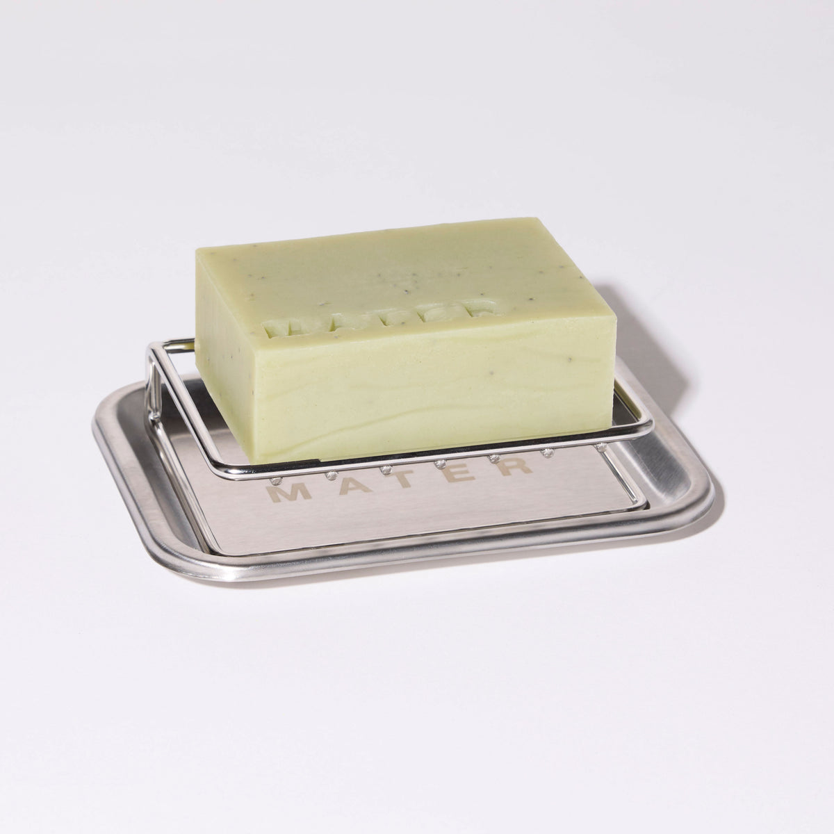 A rectangular bar of pale yellow-green soap with small dark flecks, resting on a silver metal soap dish with the brand name 'MATER' embossed on the front, set against a plain white background.