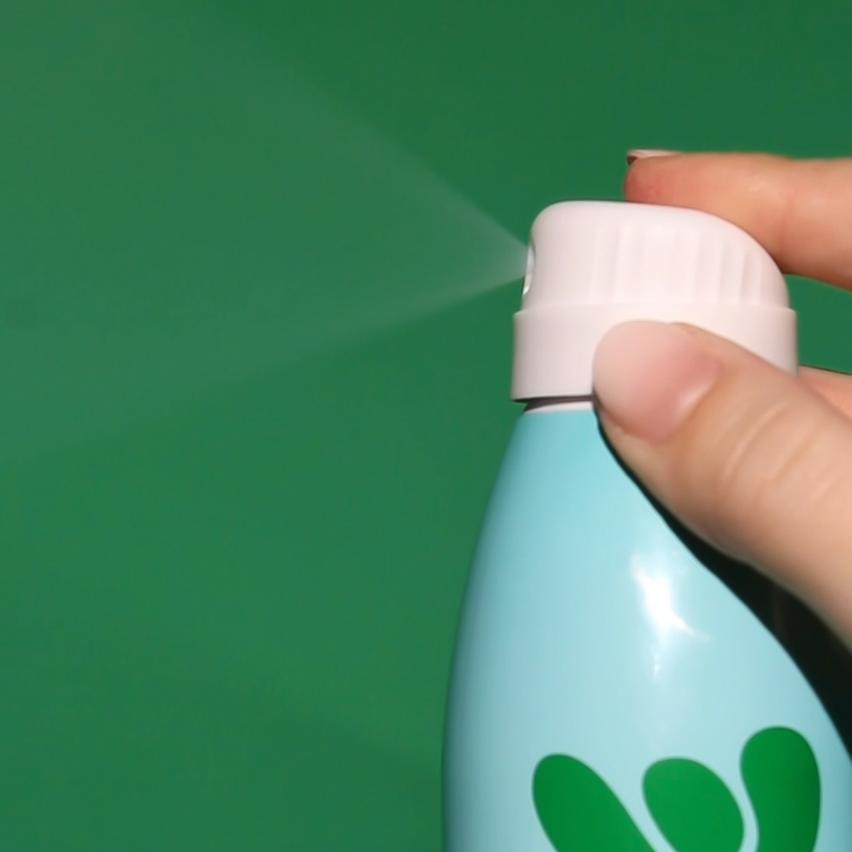 Hand holding Bask's mineral sunscreen bottle as it is spraying a fine mist in front of a green background. 