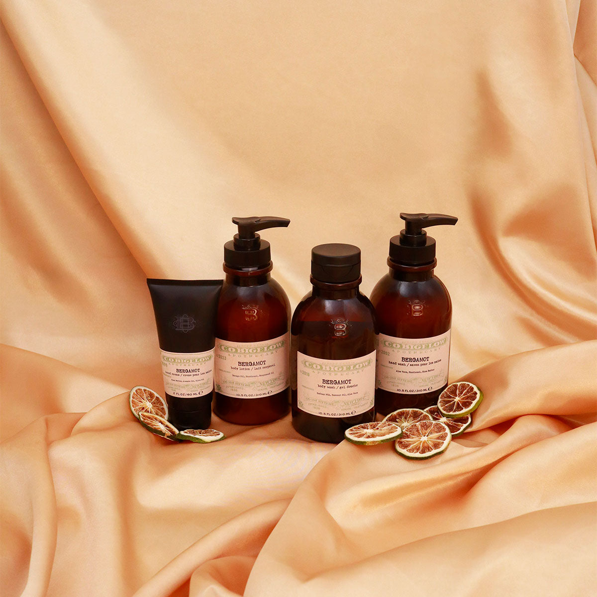 C.O. Bigelow Iconic Collection Bergamot Hand Cream, Body Lotion, Body Wash, and Hand Wash next to Bergamot slices on top of draped silk. 