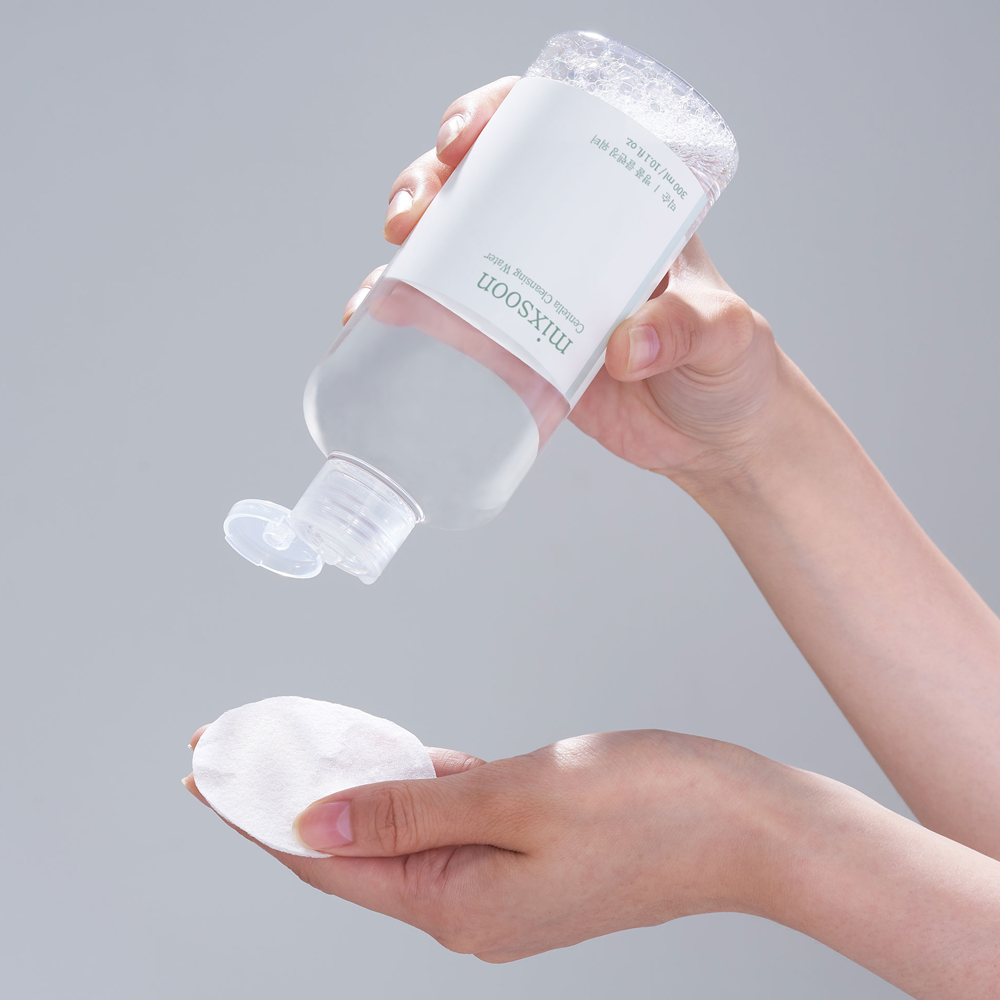 A person holds a bottle of mixsoon Centella Cleansing Water above their hand, pouring the liquid while holding a white cotton pad below. The background is a solid light gray studio backdrop.