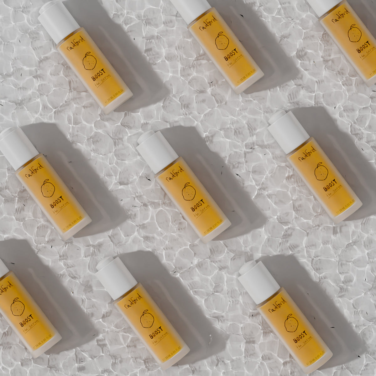 Bottles of Radford BOOST-the serum with white caps and yellow-orange liquid inside are arranged diagonally on a textured white surface. 