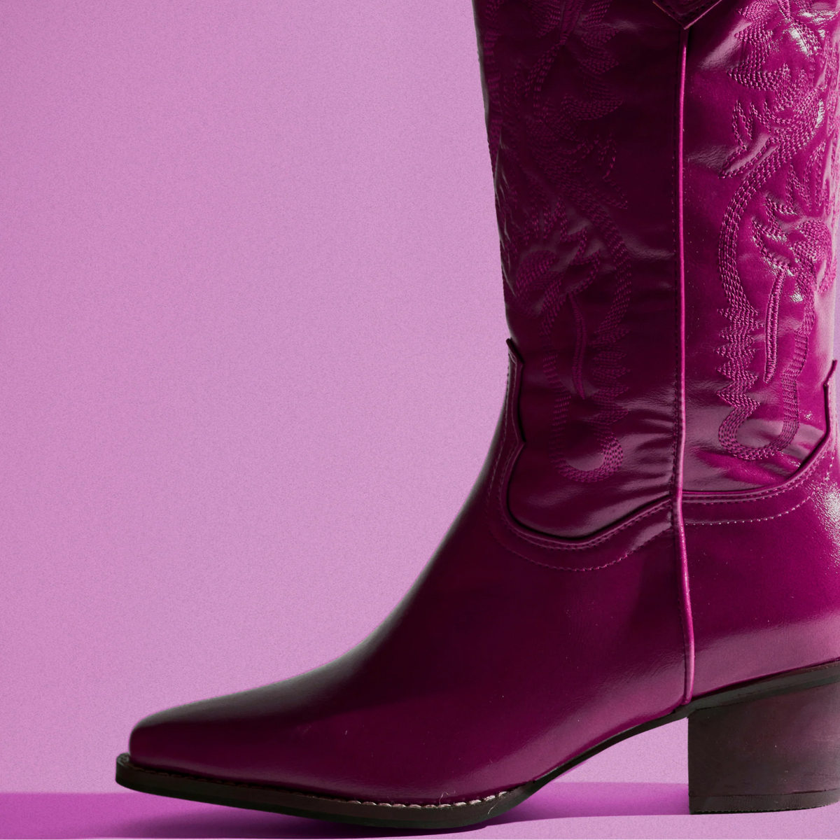 A single glossy magenta western boot with embossed floral detailing on the shaft, shown against a solid lavender-pink background.