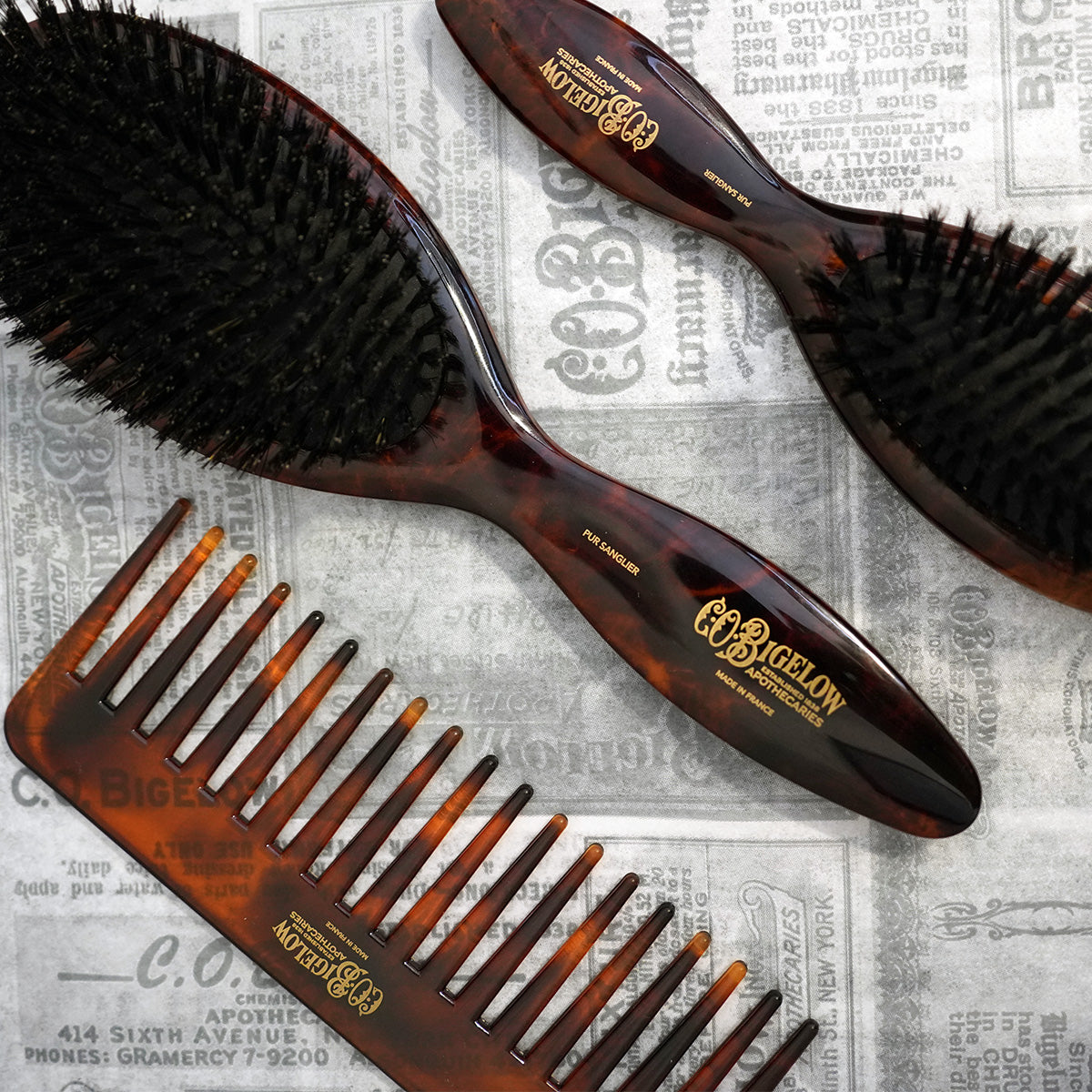 Two tortoiseshell hairbrushes and a comb on a newspaper background with 'C.O. Bigelow' branding.