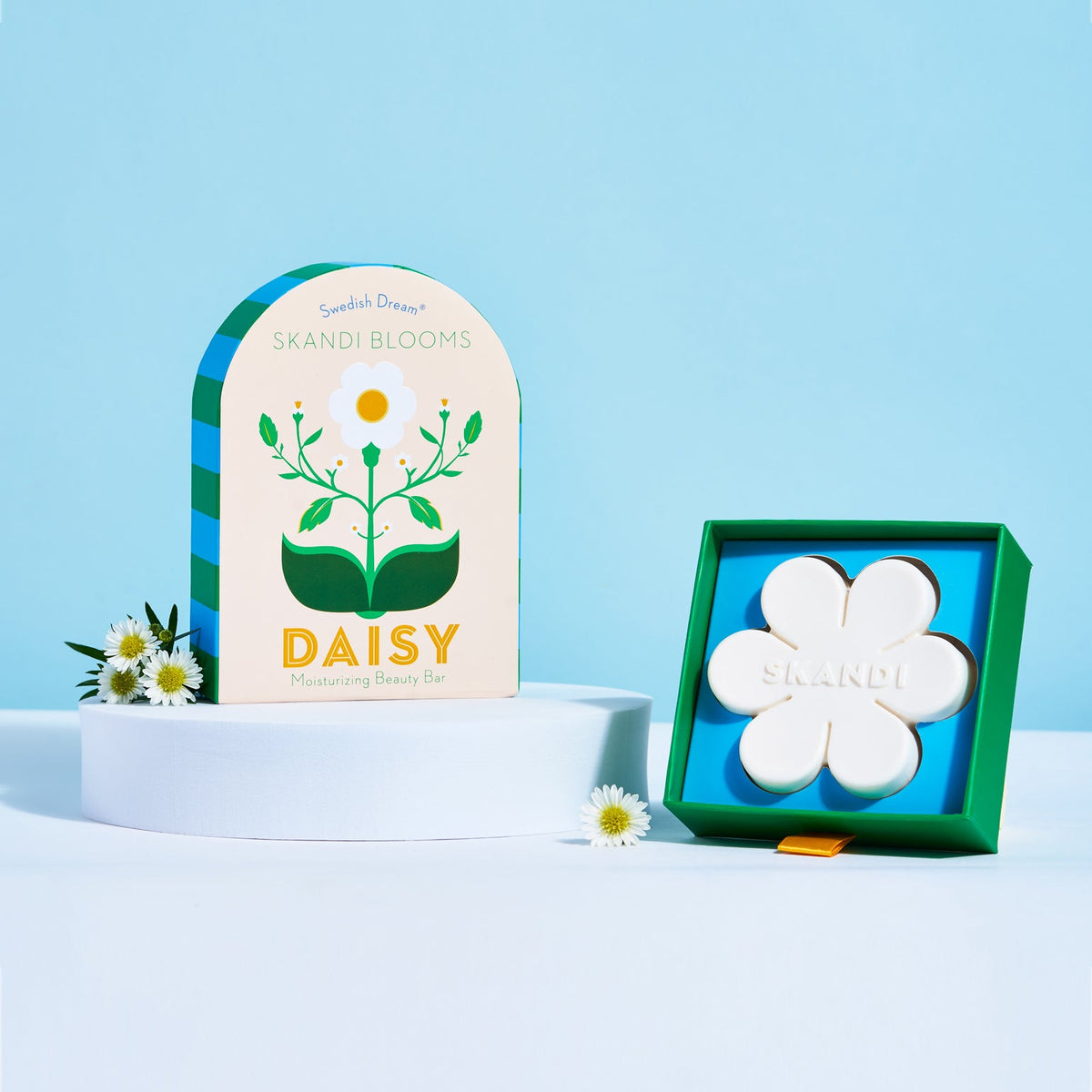 Kala Style Skandi Blooms Daisy Moisturizing Beauty Bar next to outer packaging box and fresh daisies in front of blue background.