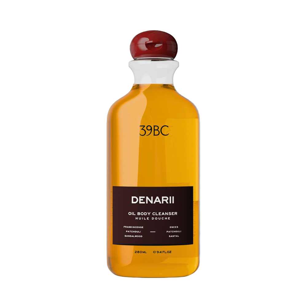 A bottle of 39BC Denarii Oil Body Cleanser with a clear glass body showing the golden-yellow liquid inside, a dark brown label, and a dark red cap, set against a plain white background.