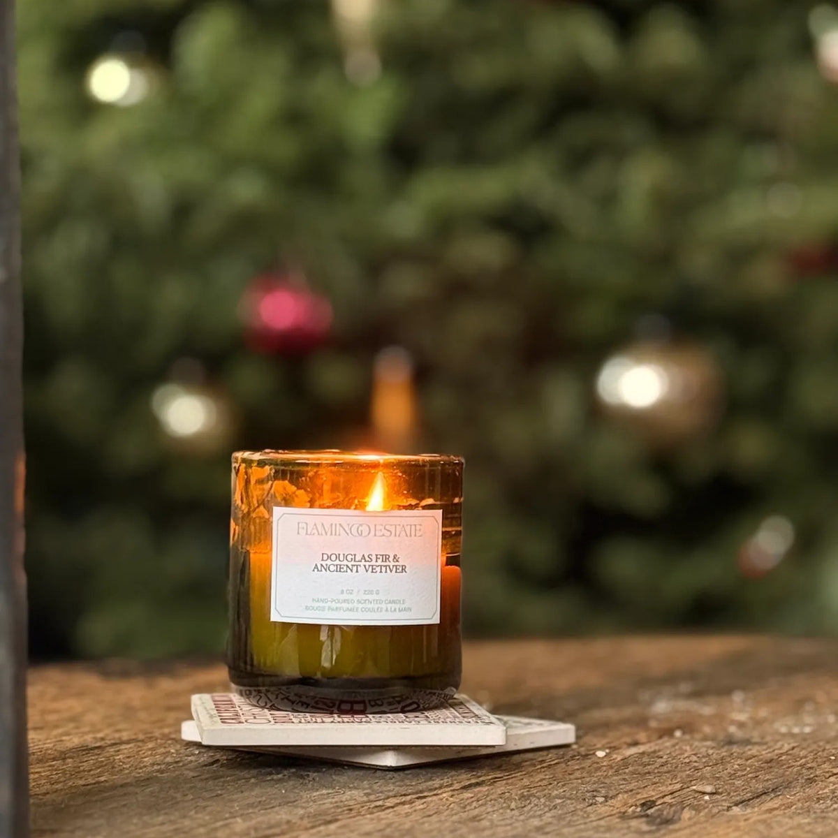 Flamingo Estate Candle with a label on a coaster in front of a blurred Christmas tree