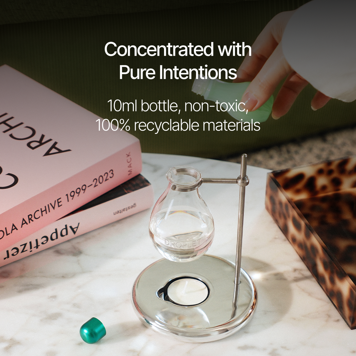 A clear glass perfume bottle with a silver metal atomizer stand is shown on a marble surface, with a hand holding the spray nozzle. White text overlays read 'Concentrated with Pure Intentions', '10ml bottle, non-toxic', and '100% recyclable materials'.