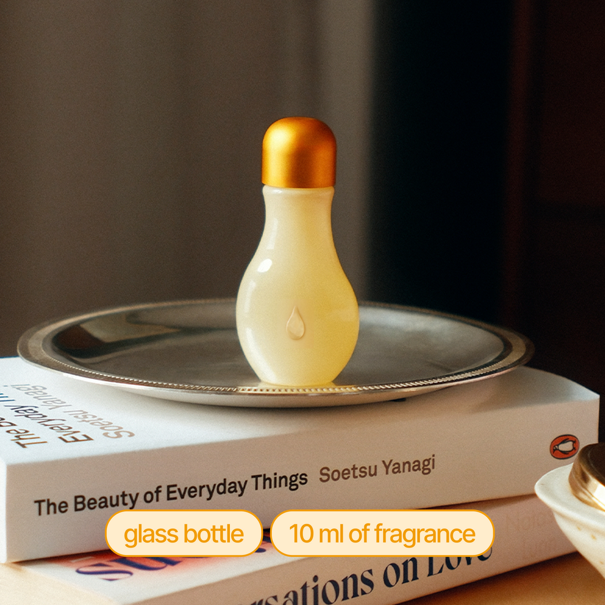 A small glass fragrance bottle with a frosted pale yellow body and a metallic gold cap, displayed on a silver tray resting on stacked books.