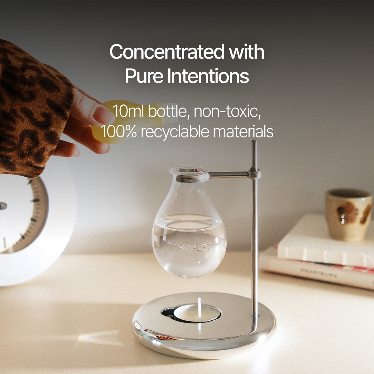 A 10ml droplet water bottle suspended on a chrome stand on a tabletop, with a hand in a leopard-print sleeve reaching toward it. White text reads 'Concentrated with Pure Intentions' and '10ml bottle, non-toxic, 100% recyclable materials.