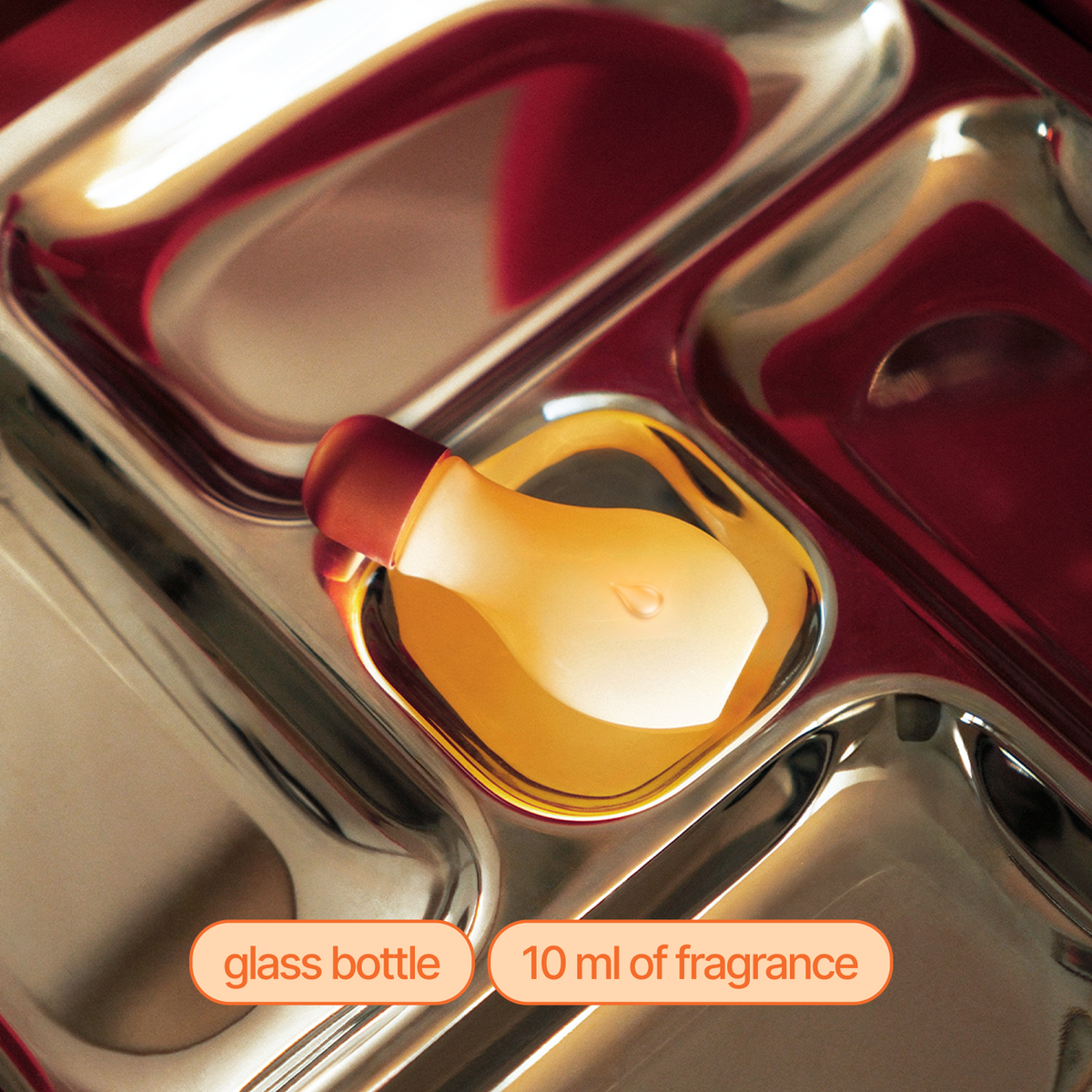 A small glass fragrance bottle with a translucent amber-yellow body and a dark red cap, resting in a metallic compartment. Text labels at the bottom read 'glass bottle' and '10 ml of fragrance'.