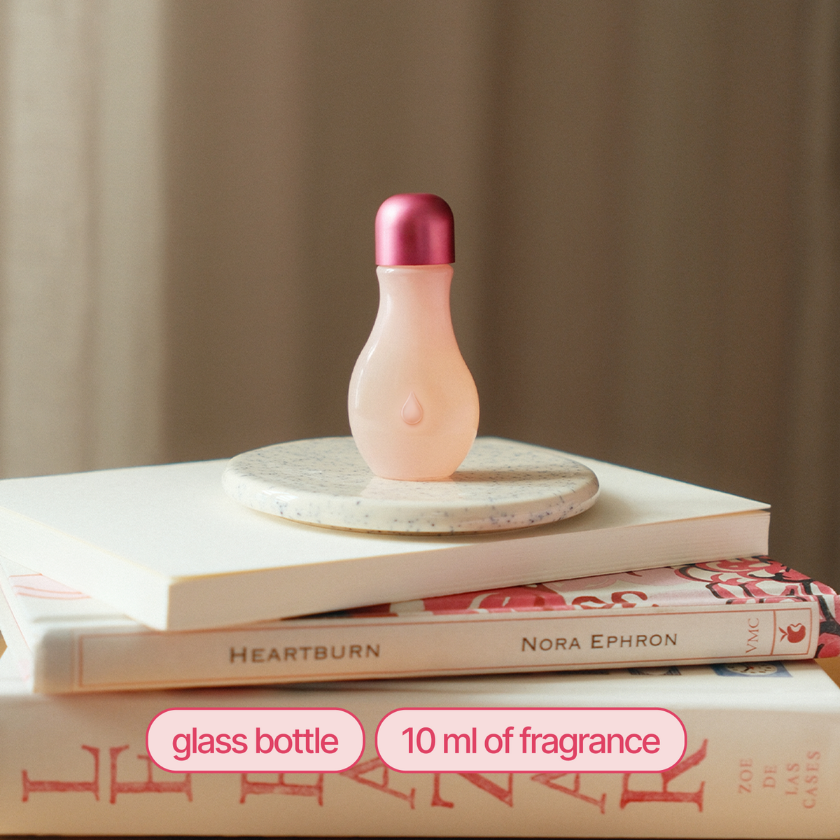A small pear-shaped glass fragrance bottle with a pale blush pink body and metallic rose gold cap, displayed on a stack of books with a marble coaster underneath. Text overlays at the bottom read 'glass bottle' and '10 ml of fragrance'.