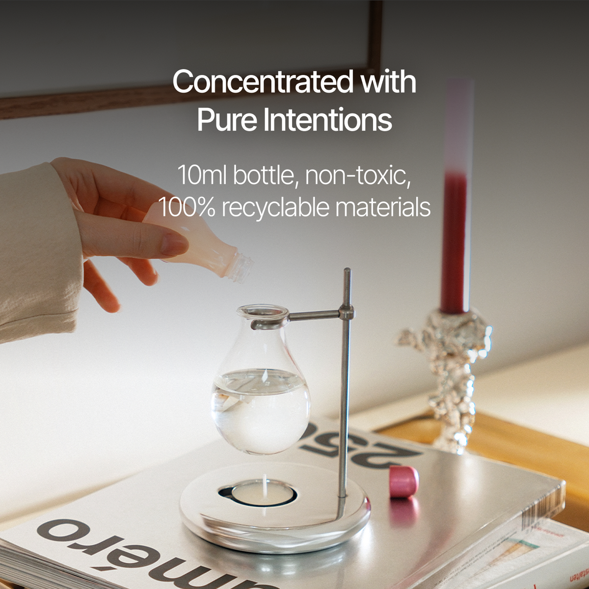 A hand holds a small clear bottle above a glass round-bottom flask on a metal stand, with overlaid text reading 'Concentrated with Pure Intentions' and '10ml bottle, non-toxic, 100% recyclable materials.' The scene is styled on a tabletop with a candle and magazine in the background.