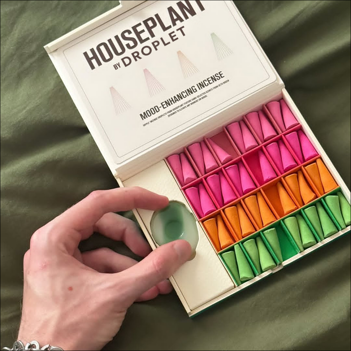 A hand holds a small green droplet-shaped incense piece beside an open white box labeled 'HOUSEPLANT BY DROPLET' containing multiple colorful incense pieces in pink, orange, and green, set against an olive green fabric background.