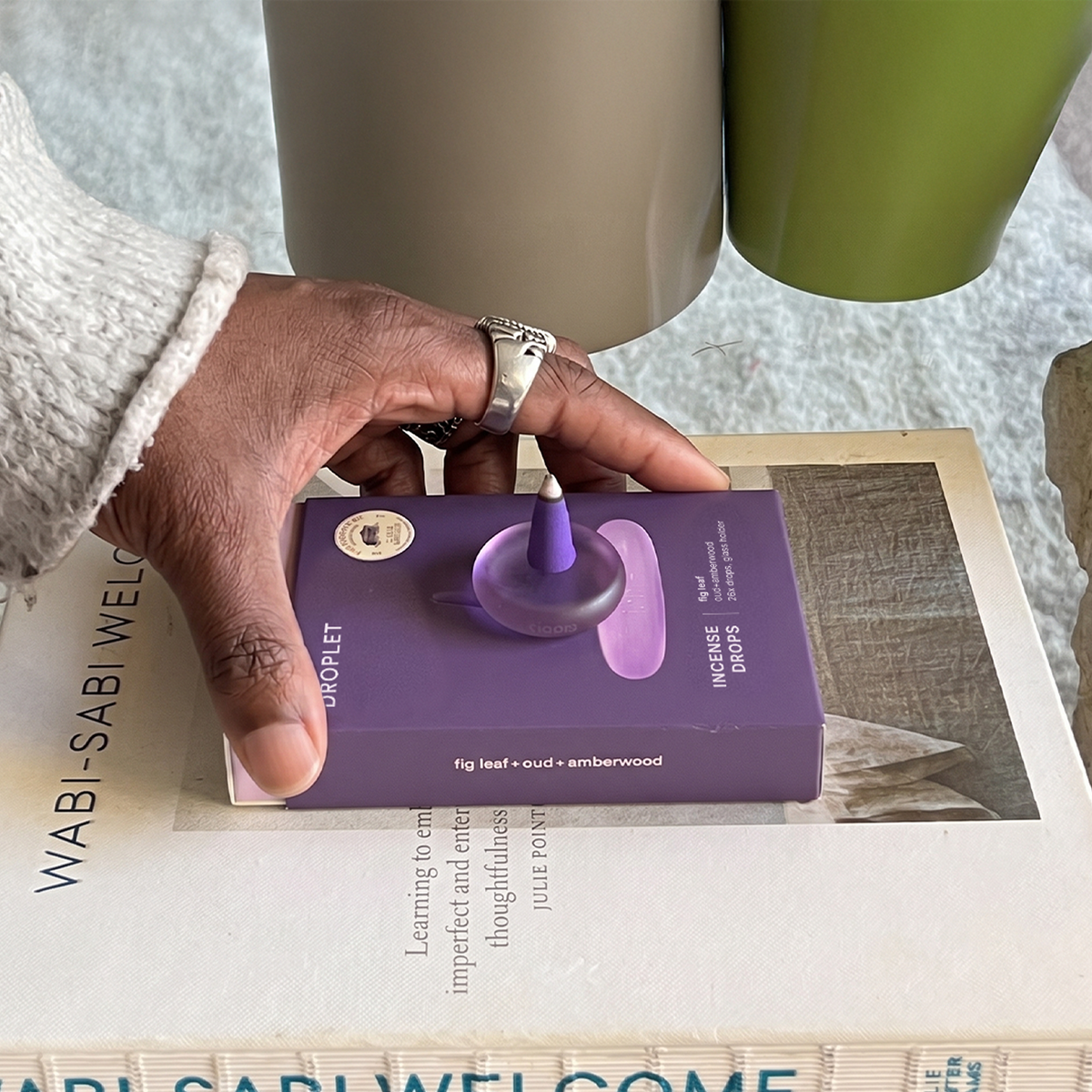 A hand wearing a white knit sweater and a silver ring holds a purple box of Wabi Sabi Wax Erasers over a book.