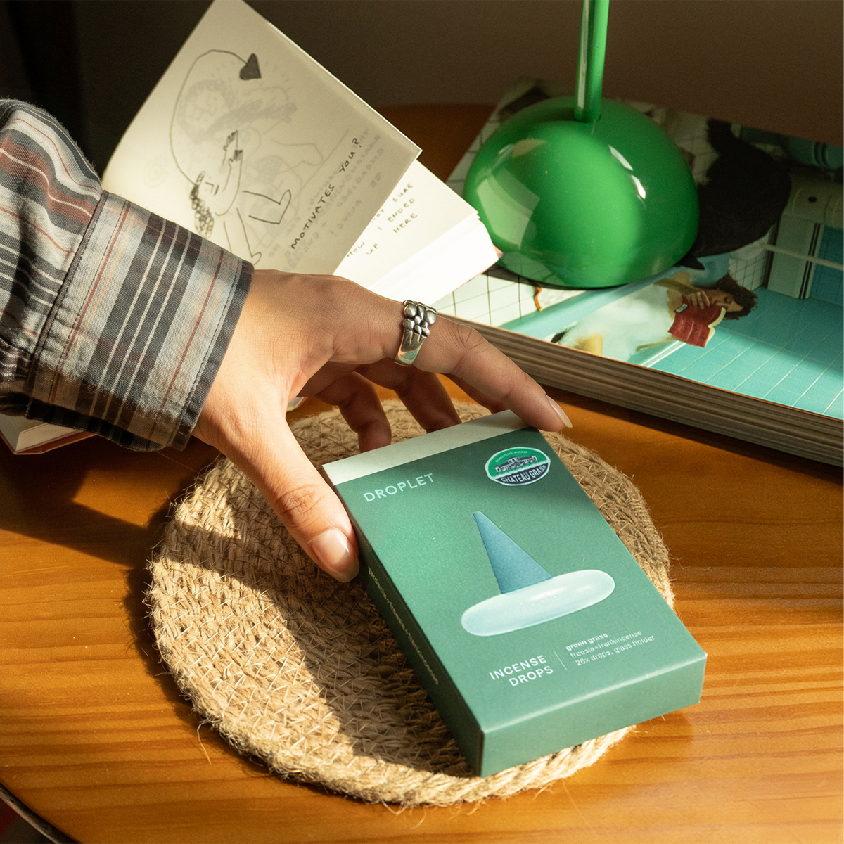 A hand wearing a plaid shirt and silver ring holds a green box of DROPLET Incense Drops on a woven placemat over a wooden table, with books and a green lamp in the background.