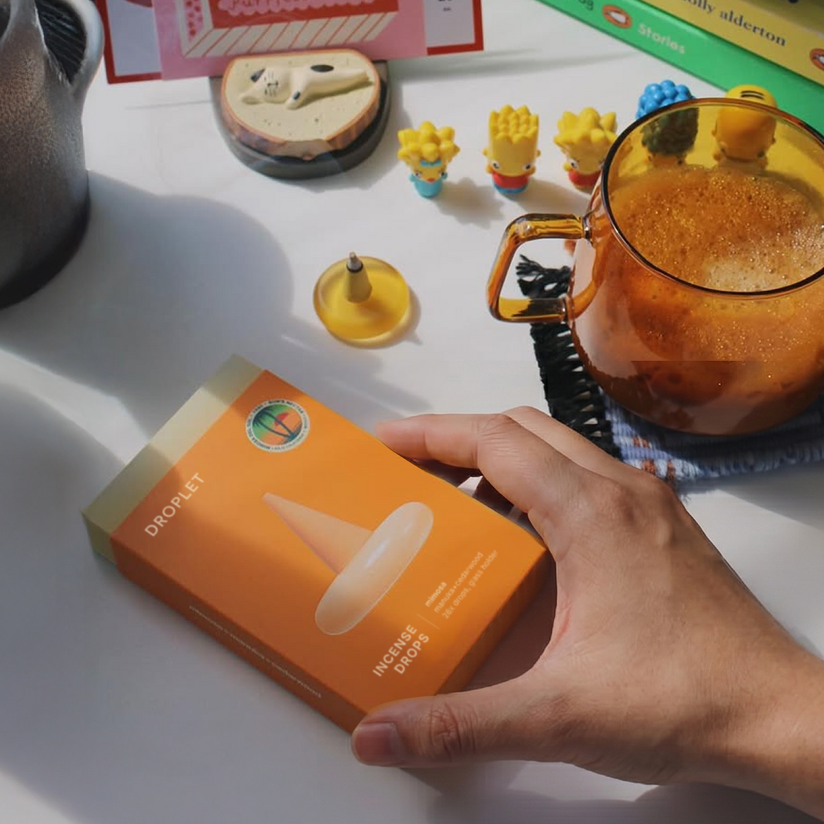 A hand holds an orange box labeled 'DROPLET Incense Drops' on a white tabletop. The scene includes a glass mug filled with amber-colored liquid, small figurines, and other desk items in warm natural light.