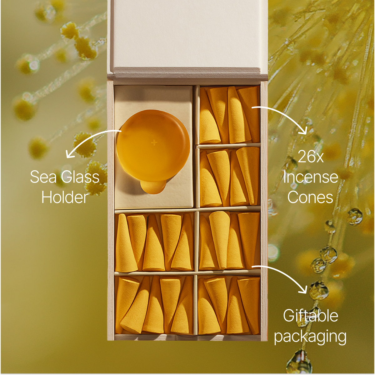 An open gift box containing 26 yellow-orange incense cones arranged in compartments, with a round amber glass holder. White callout lines and text labels identify the incense cones, sea glass holder, and giftable packaging against a soft yellow background with droplet graphics.