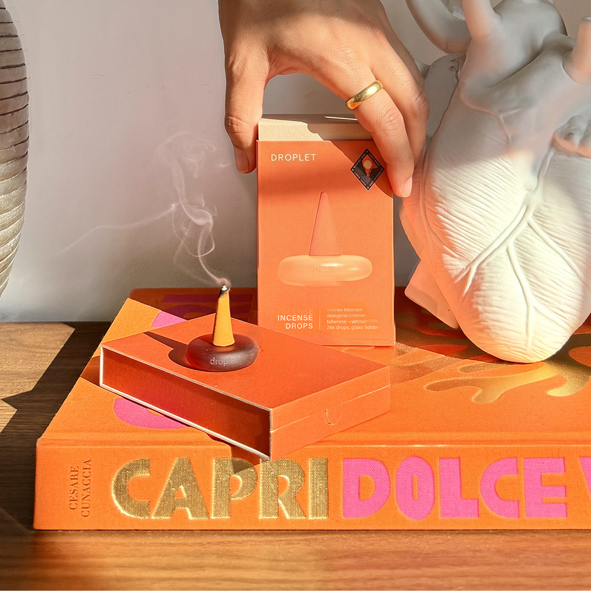 A hand holds an orange box labeled 'DROPLET INCENSE DROPS' above an open orange matchbox containing a smoking incense cone, styled on top of a large orange book with gold and pink lettering. A white anatomical heart sculpture and a woven basket are visible in the background.