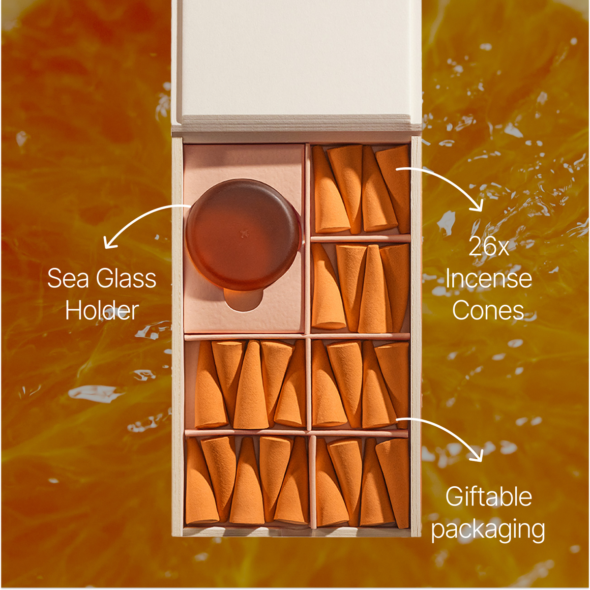 An open wooden gift box containing 26 orange incense cones arranged in compartments, with a round amber-colored sea glass holder in one slot. White callout lines and text labels identify the sea glass holder, 26 incense cones, and giftable packaging against an orange textured background.