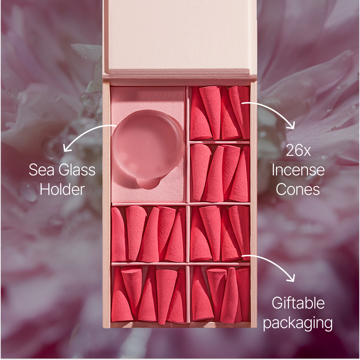 An open pink gift box containing 26 pink incense cones arranged in compartments, with a round translucent pink sea glass holder in one slot. White callout lines and text label the sea glass holder, incense cones, and giftable packaging against a soft blurred floral background.